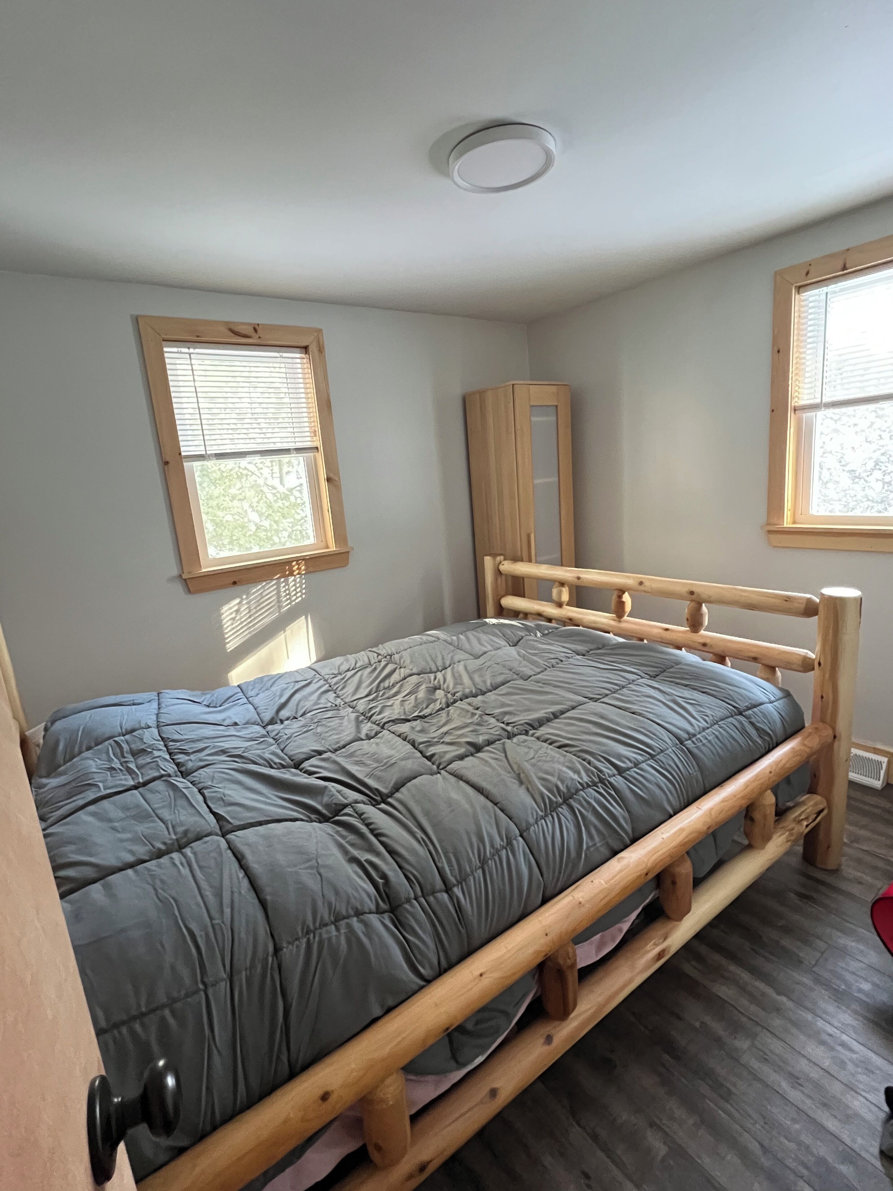 A single bed with a wooden frame and a grey quilt, a window with blinds, and a mirror on the wall.