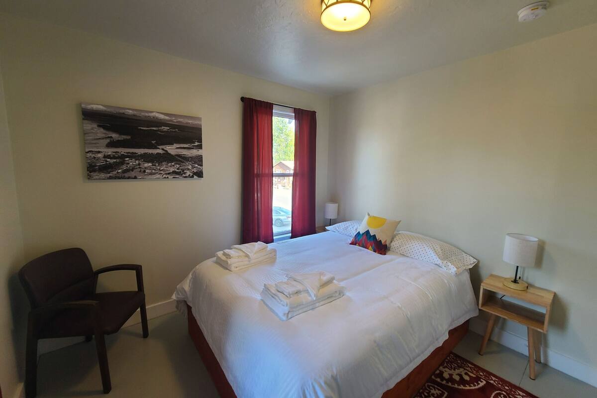 Une chambre d’hôtel avec un lit, une chaise, une table de chevet, une lampe, un tableau et une fenêtre avec des rideaux.