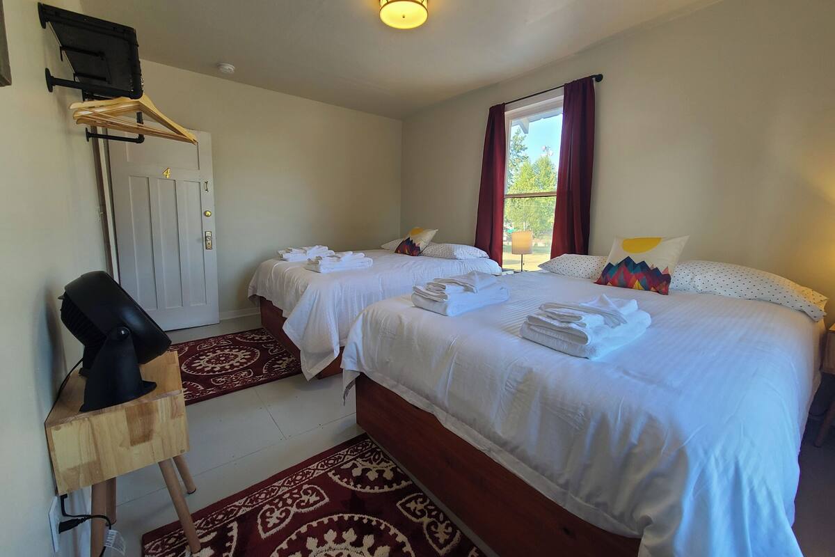 Une chambre d’hôtel avec deux lits, un sèche-cheveux posé sur un support en bois, un tapis rouge à motifs et une fenêtre avec des rideaux.