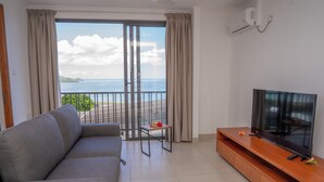 A living room with a grey sofa, a wooden TV stand, and a sliding glass door leading to a balcony with a view of the sea. - Villa Kolibri (Mahé Island)
