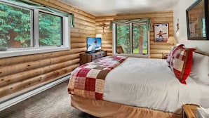 Un dormitorio tipo cabaña de madera con una cama, un televisor y una ventana con vista a árboles.