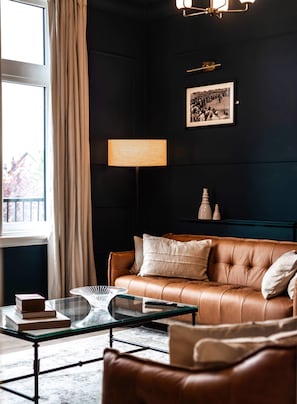 Un salon à l’ambiance sombre, avec un canapé en cuir, une table basse en verre, une lampe et un tableau encadré au mur.