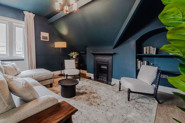 A room with a dark blue wall, a fireplace, a sofa, a chair, a round table, and a bookshelf.