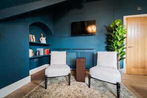 A living room with two white armchairs, a wooden coffee table, a bookshelf, a television mounted on the wall, and a large green plant.