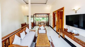 A spacious living area with a sofa, a coffee table, and a TV. There is a wooden door leading to another room and a window with a view of greenery. - Tirtodipuran Hotel  (Yogyakarta)