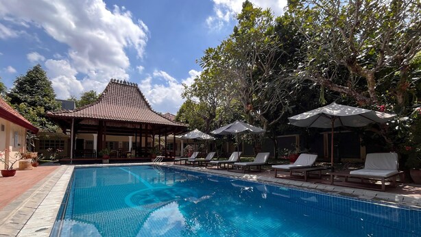 A swimming pool with lounge chairs and umbrellas, surrounded by trees and a building with a tiled roof. - Tirtodipuran Hotel  (Yogyakarta)