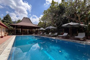 Ein Pool mit Liegestühlen und Sonnenschirmen, umgeben von Bäumen und einem Gebäude mit einem Ziegeldach.