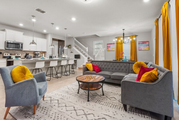 A modern living room with a grey sofa, two armchairs, a coffee table, and a kitchen area in the background. - Disney Family Escape w/ Pool, Game Room + Themed Rooms (Davenport)