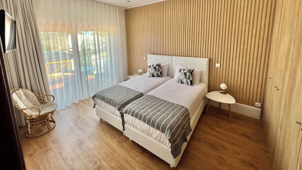 A double bed with a striped bedspread, a bedside table with a lamp, and a view of the outdoors through large windows with sheer curtains. - THE CORNER OF THE RIA (Torreira)