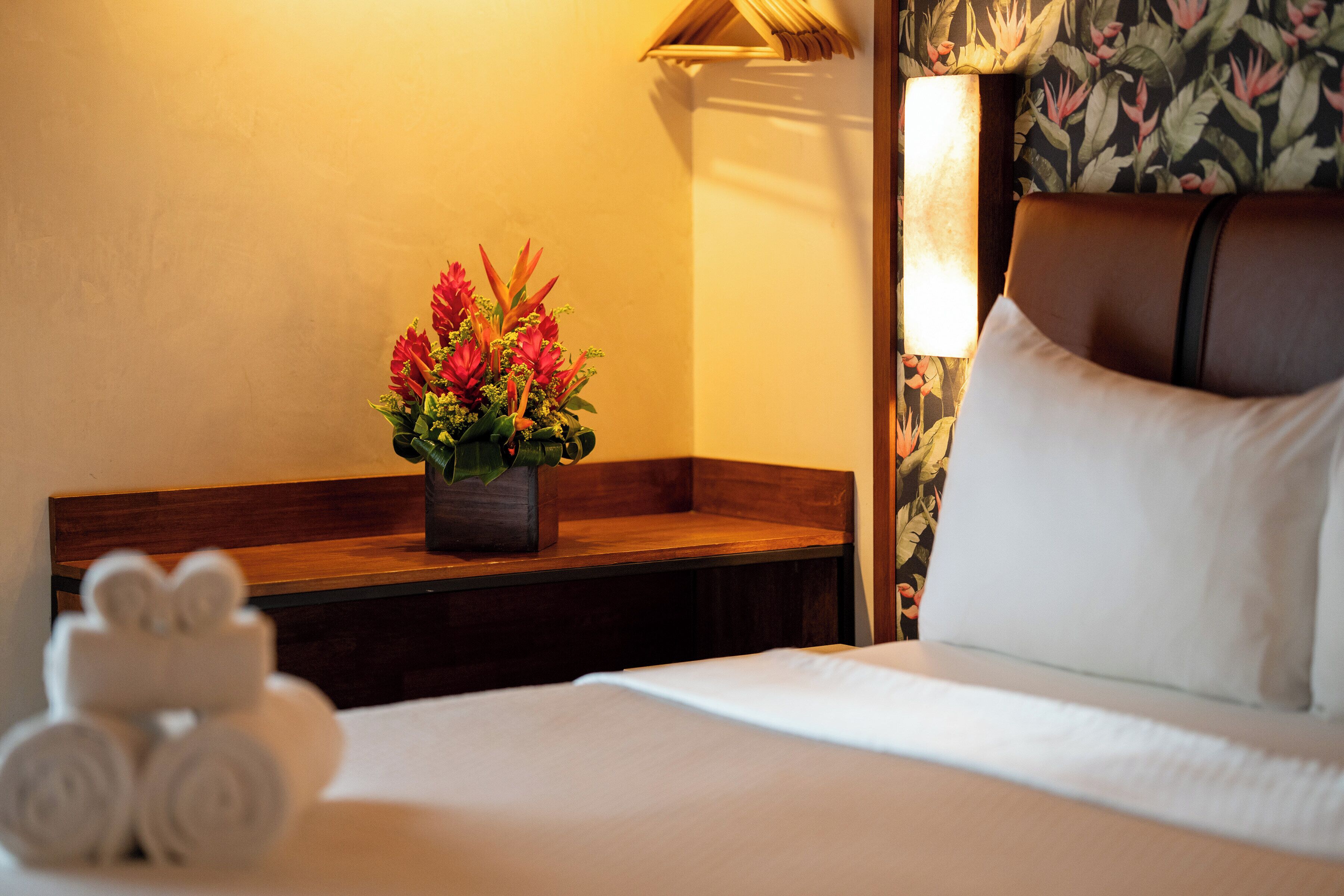 Ein Hotelzimmer mit einem ordentlich bezogenen Bett, einem Nachttisch aus Holz mit einer Lampe mit Blumenmuster und einer Zierpflanze.