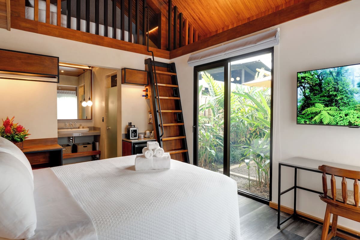 A bedroom with a large bed, a wooden staircase, a bathroom, and a sliding glass door leading to a garden.