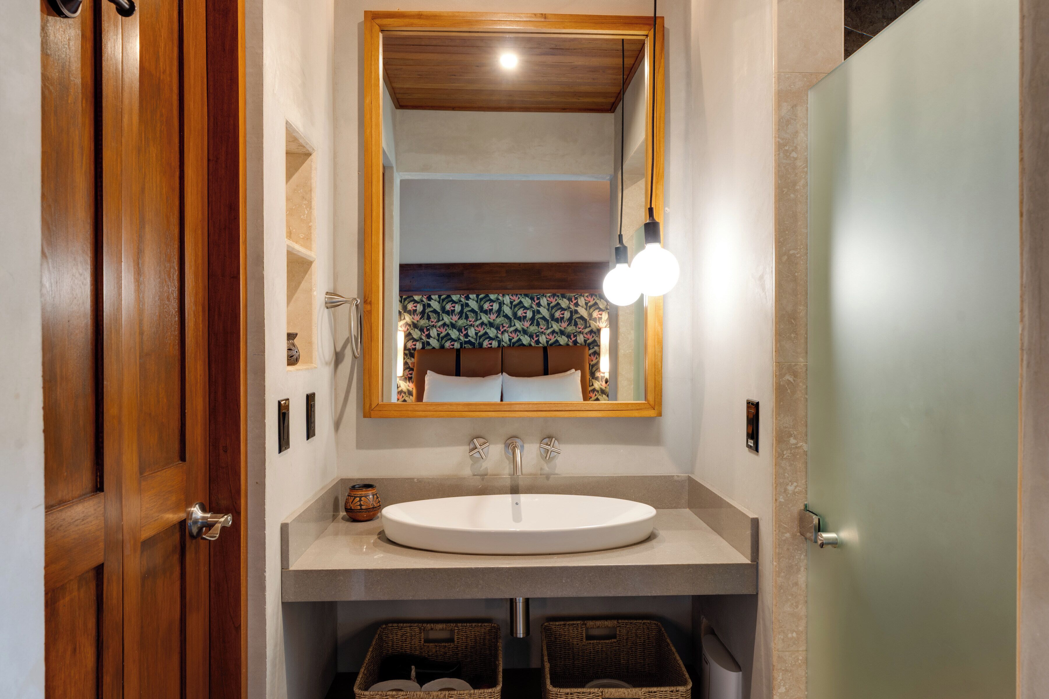 Un baño con puerta de madera, lavamanos blanco, espejo con luces y cestos de mimbre debajo del lavamanos.