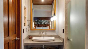 Un baño con puerta de madera, lavamanos blanco, espejo con luces y cestos de mimbre debajo del lavamanos.