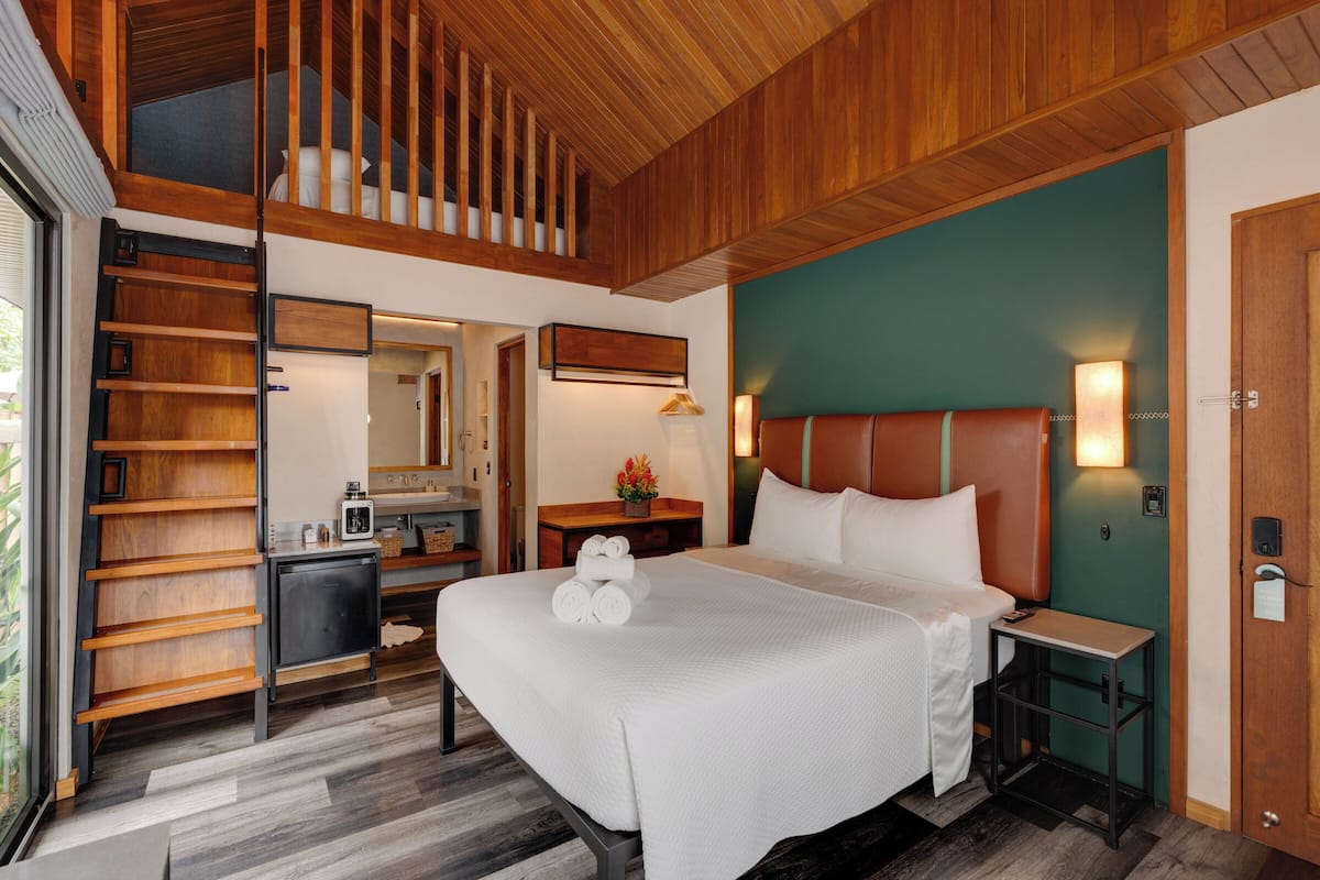 A hotel room with a bed, a desk, a small kitchenette, a mirror, and a staircase.