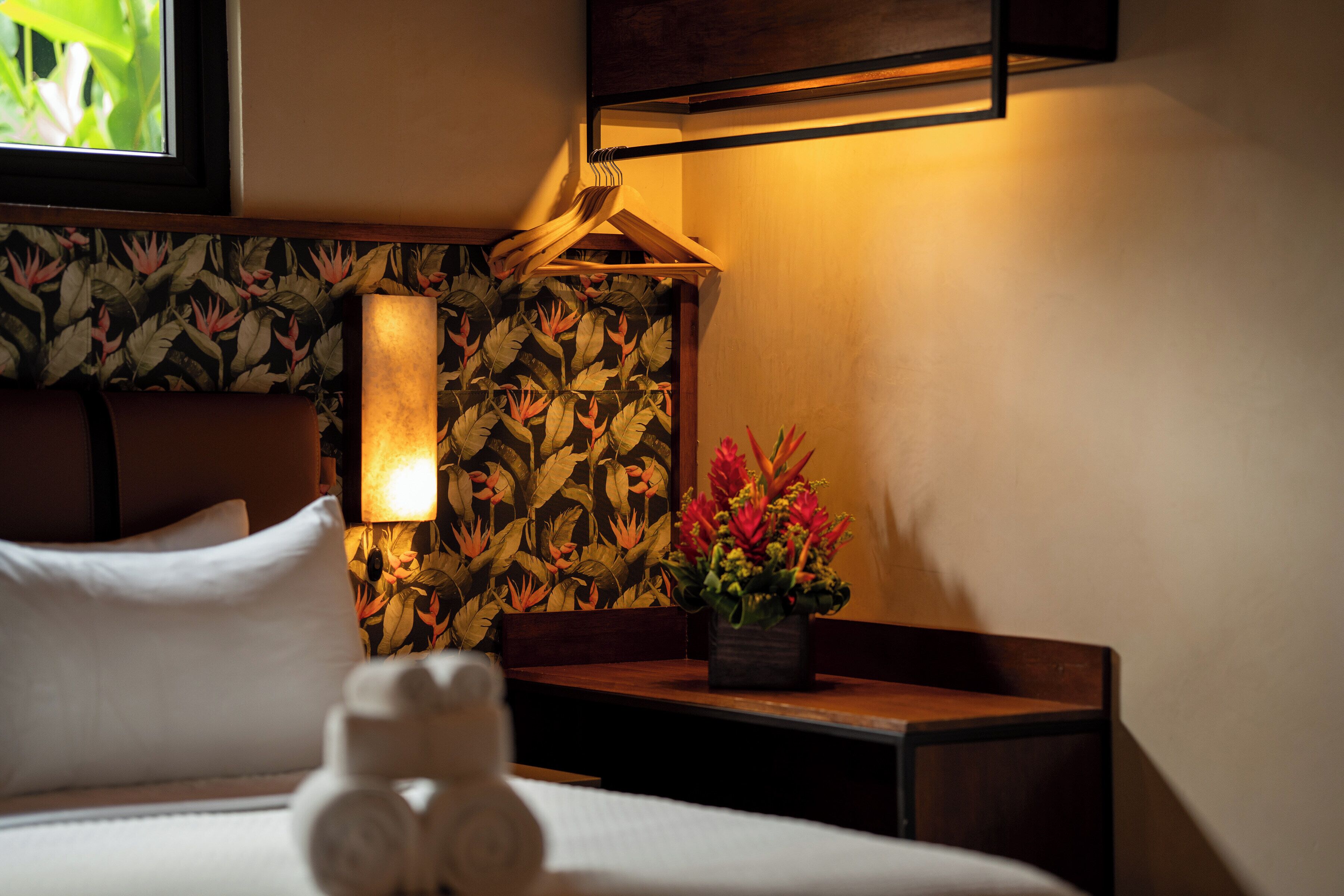 A hotel room with a bed, a nightstand with a lamp, a floral wallpaper, and a small table with a flower vase.