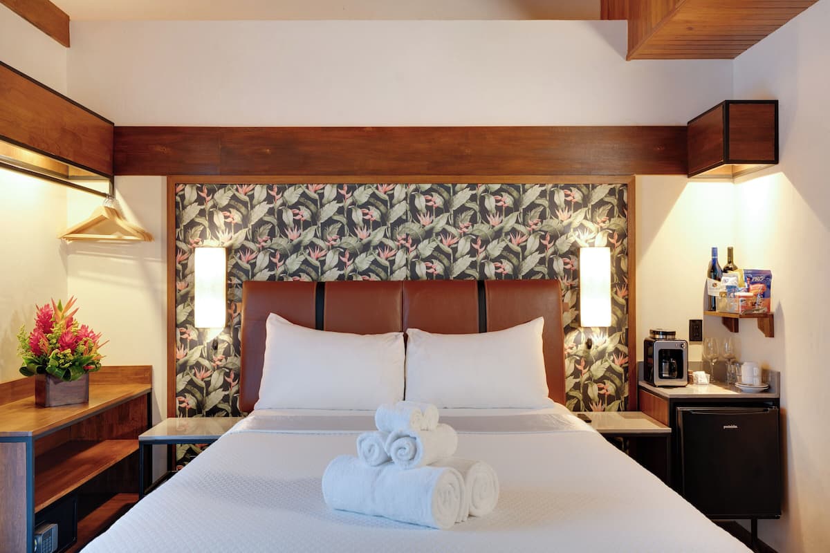 A hotel room with a bed, a small desk, a refrigerator, and a nightstand with a lamp.