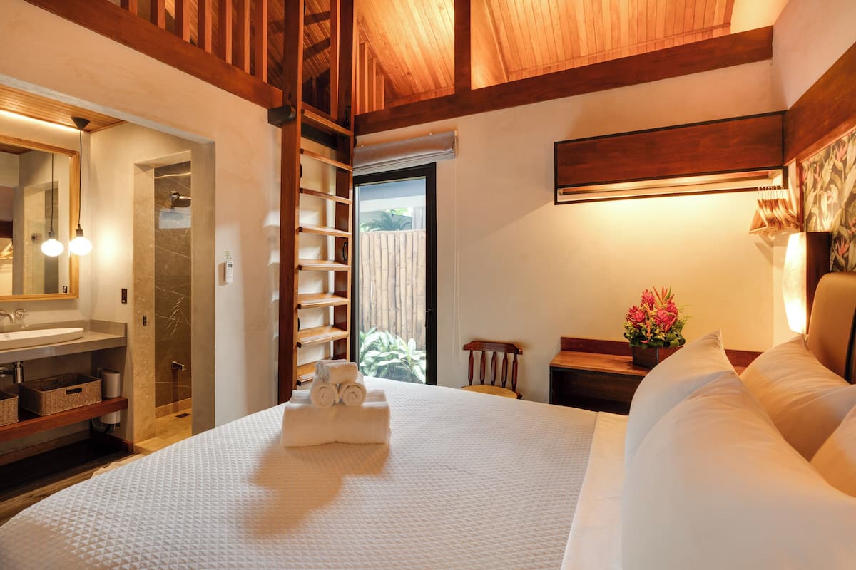 A well-lit bedroom with a large bed, a wooden headboard, a bedside table with a lamp, and a view of the outdoors through a window.