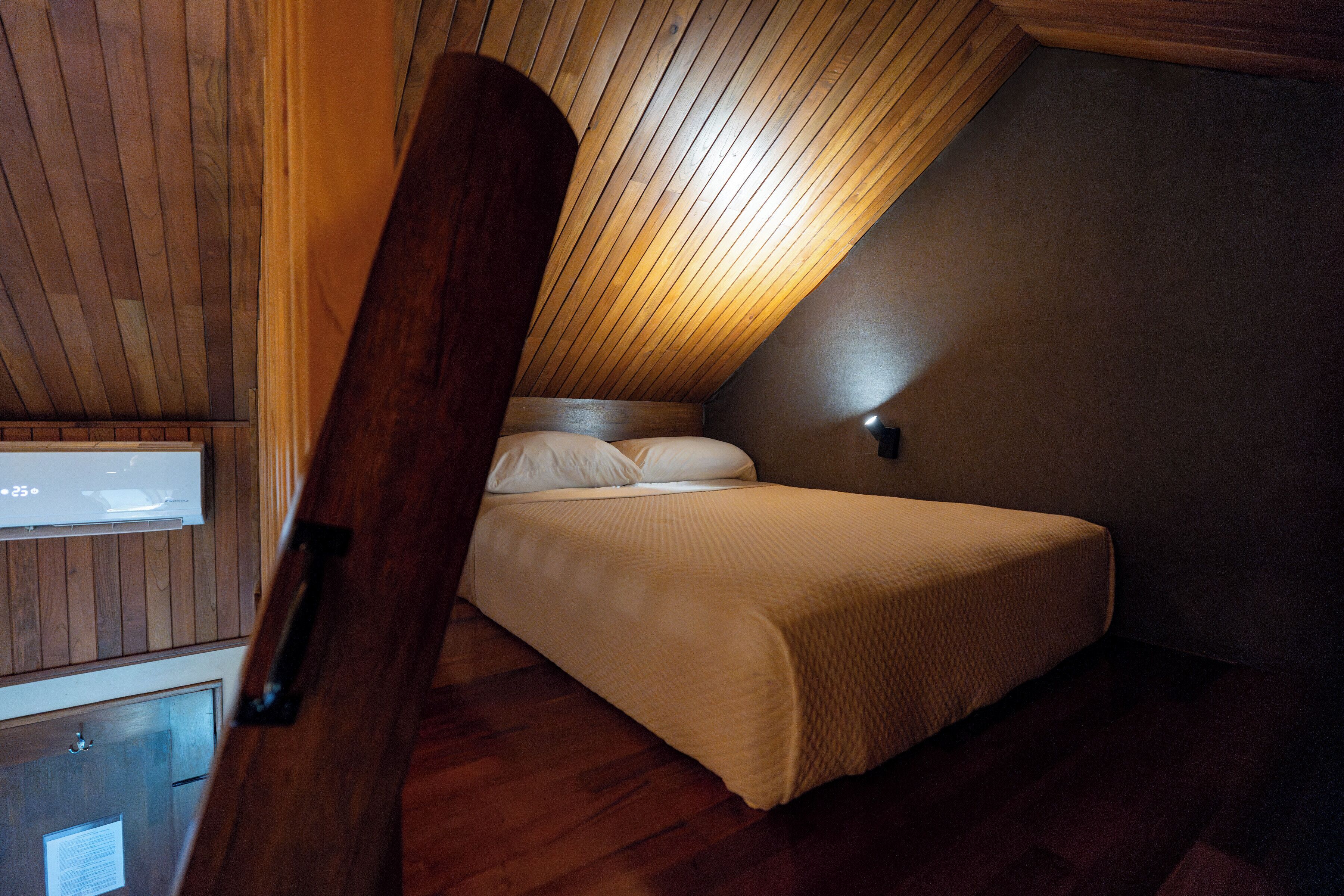 Ein Schlafzimmer mit geneigter Decke, Holzwänden, einem Bett mit weißer Bettwäsche und einem kleinen, an der Wand befestigten Fernseher.