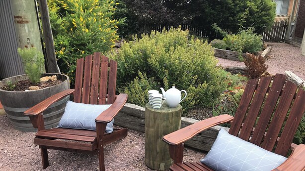 Two wooden Adirondack chairs with cushions, placed on a gravel surface in a garden setting with various plants and a wooden post with a teapot and cups. - 
Air's Cottage Penola: Colonial stone cottage, WiFi fire AC Spa BBQ dog friendly (Penola)