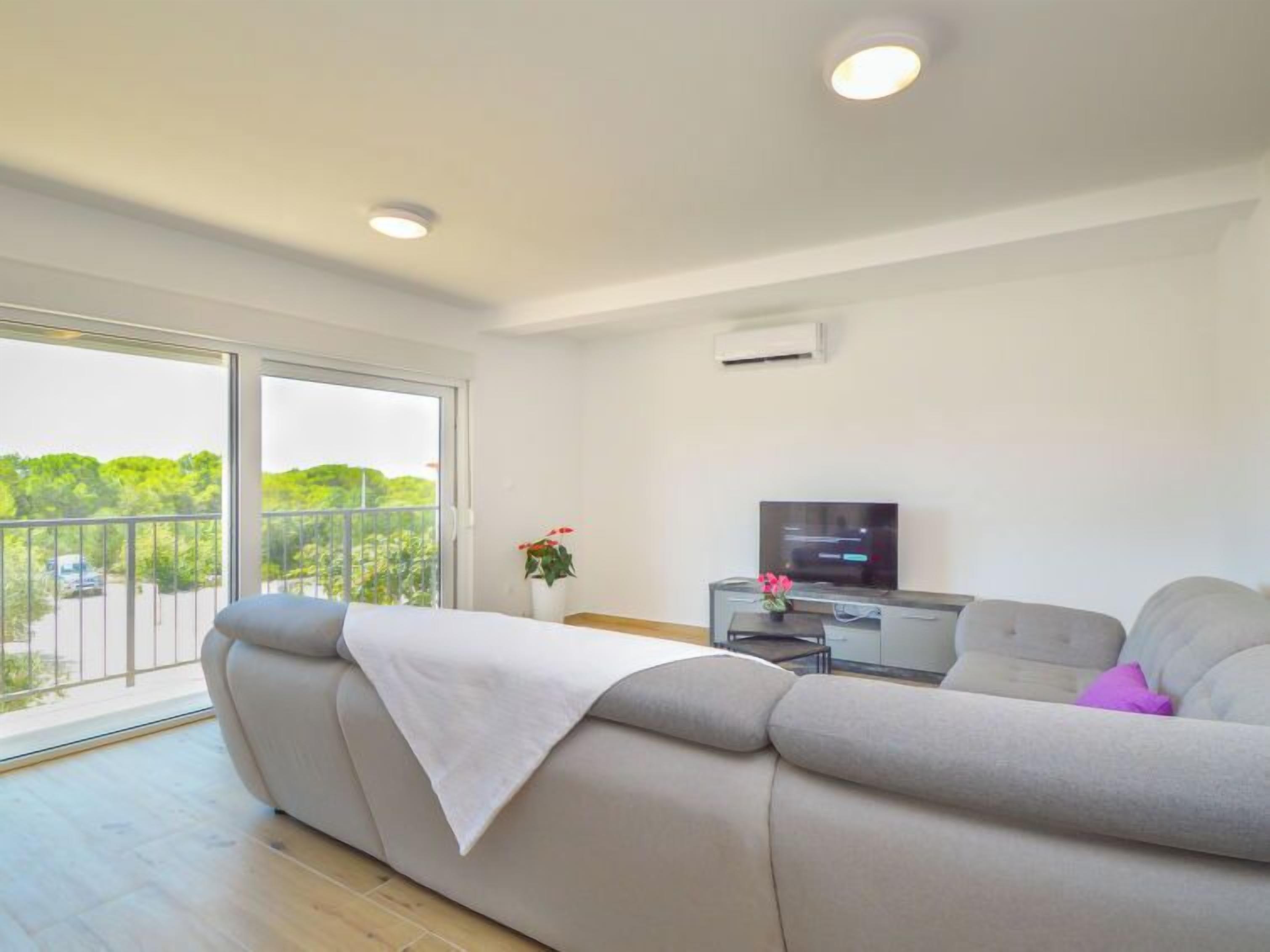 A modern living room with a sofa, a TV, and a balcony with a view of greenery.