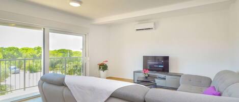 A modern living room with a sofa, a TV, and a balcony with a view of greenery.