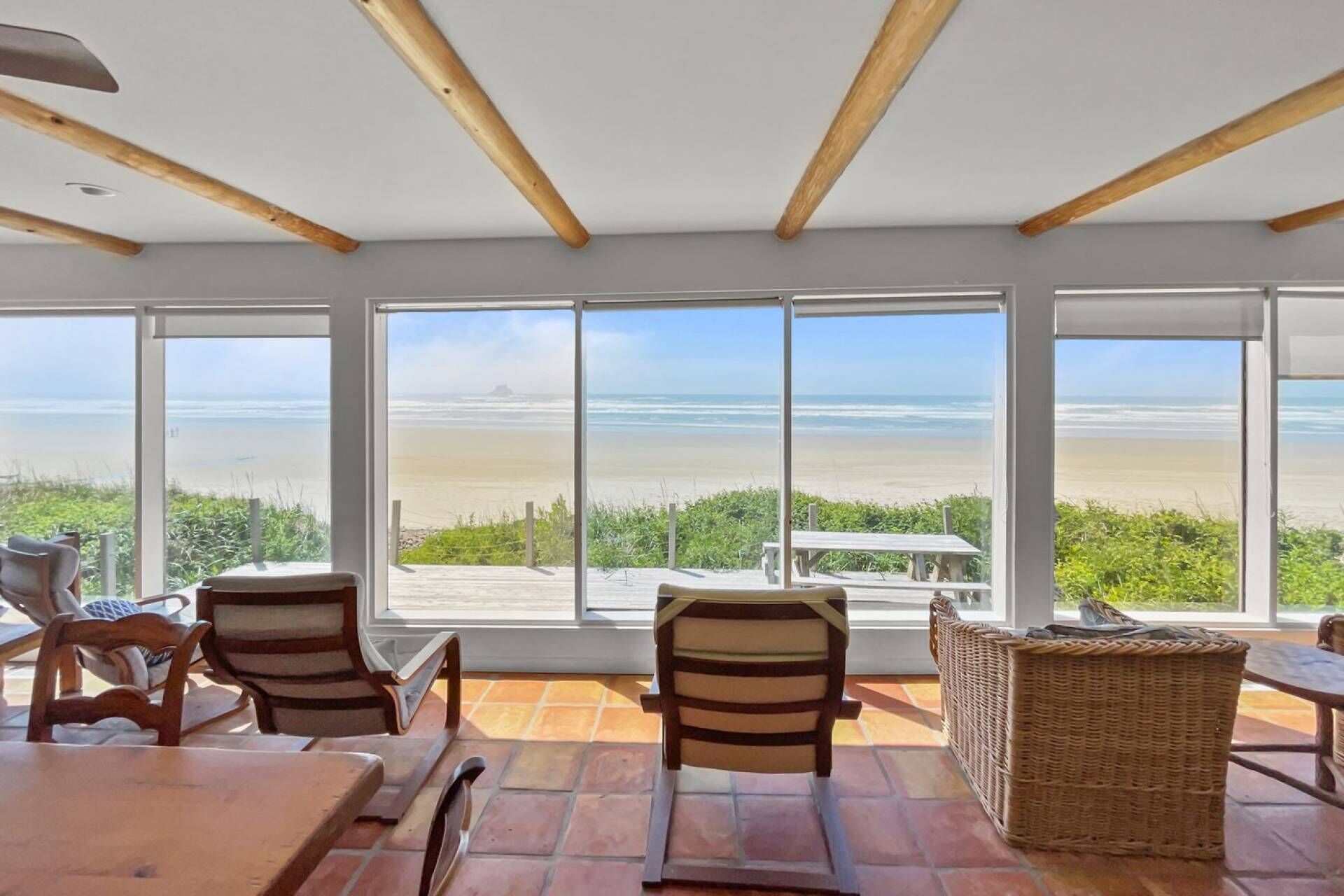 A beachside room with large windows, wooden ceiling beams, and a wicker table.