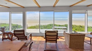 A beachside room with large windows, wooden ceiling beams, and a wicker table. - Coastal Escape with Stunning Views, Pet-Friendly Comfort, Hot Tub & Deck Just Steps to the Sand (Arch Cape)