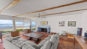 A spacious living room with a large gray sofa, a wooden coffee table, and a fireplace. There are large windows offering a view of the beach. - Coastal Escape with Stunning Views, Pet-Friendly Comfort, Hot Tub & Deck Just Steps to the Sand (Arch Cape)