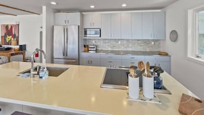 A modern kitchen with grey cabinets, a stainless steel refrigerator, a microwave, and a large island with a sink and utensils. - Coastal Escape with Stunning Views, Pet-Friendly Comfort, Hot Tub & Deck Just Steps to the Sand (Arch Cape)