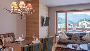 Uma sala de estar moderna com área de jantar, um lustre, um sofá com vista e uma televisão na parede.