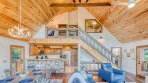 A spacious interior with a high ceiling, wooden beams, a dining area, a living room with blue furniture, and a staircase leading to an upper level. - Pineview Escape - Hot tub, arcade & mountain views (Blue Ridge Mountains)