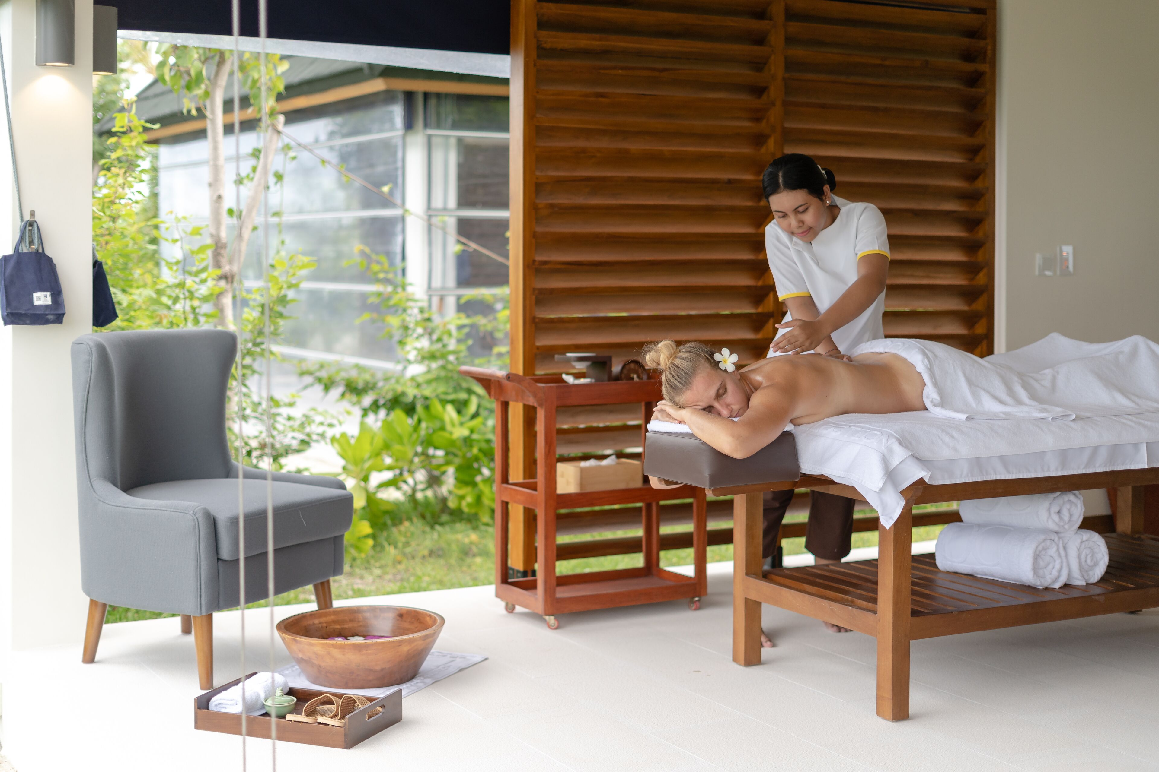 A massage therapy room with a wooden massage table, a massage table, a grey chair, a wooden side table, and a window with a view of greenery.