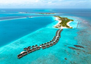 Estructura de bungalós sobre el agua con un paseo continuo, rodeada de aguas turquesas cristalinas y una pequeña isla con edificios.