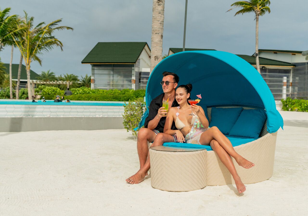 A couple sitting on a beach chair with a blue canopy, a pool, and palm trees in the background.