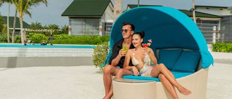 A couple sitting on a beach chair with a blue canopy, a pool, and palm trees in the background.