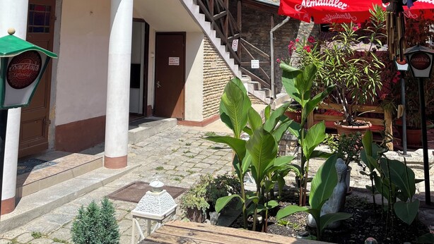 A building with a wooden staircase leading to a balcony, a stone wall, and a clock tower in the background. - Győr (Gyor)