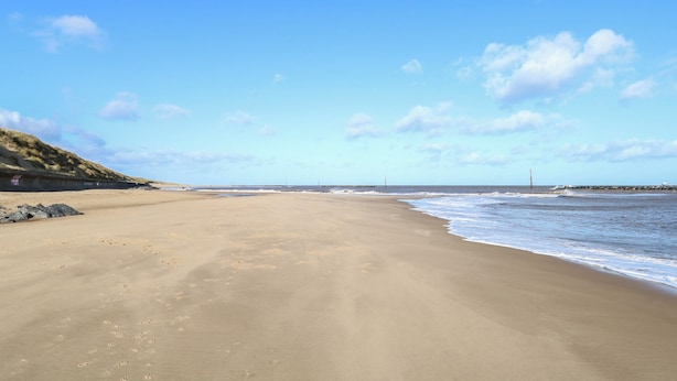 A wide sandy beach with gentle waves, a clear sky, and a rocky outcrop on the left. - The Look Out (Norwich)