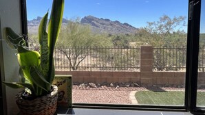 A view from a window with a potted plant, a brick wall, and a fenced area with trees and mountains in the background. - DesertOasis@ #LambertPark:4BD, Mtn Views, Gym, FREE Heated HOA Pool&Spa, PoolTable, CottonBedding (Tucson)