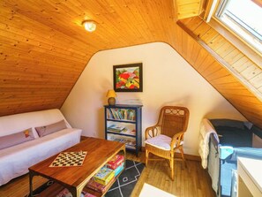 Quarto de sótão aconchegante com teto de madeira, um sofá branco, estante de livros, cadeira de vime e uma pequena mesa com um tabuleiro de xadrez.