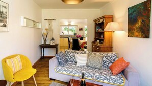Ein Wohnzimmer mit einer Couch, einem gelben Sessel, einem Essbereich mit Tisch und Stühlen sowie einer Küche im Hintergrund.