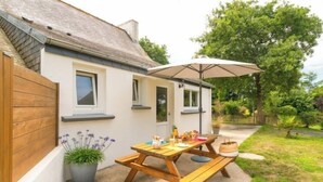 Ein kleines Haus mit einer Terrasse, auf der sich ein Holztisch und eine Bank, ein Blumentopf und ein großer Sonnenschirm befinden.
