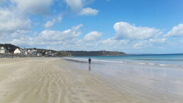 A wide sandy beach with a person walking along the shore, a clear blue sky, and houses in the background. - L'Appart'Mer by Interhome (Plérin)