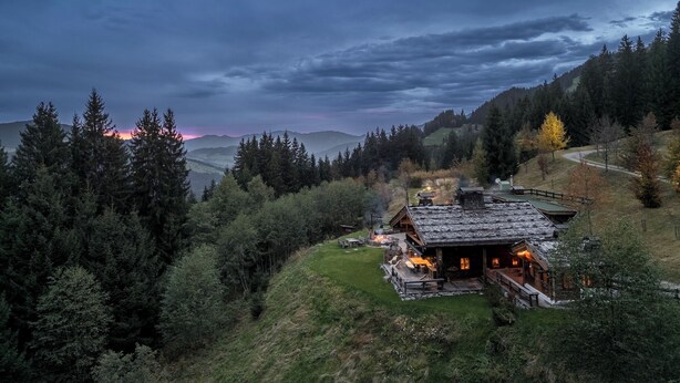 Una cabaña rústica con techo de piedra, rodeada de un bosque frondoso y vistas a montañas distantes.