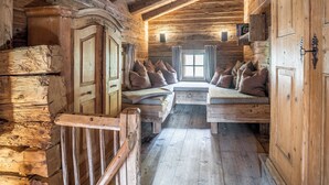 Interior de una cabaña de troncos con una cama en el altillo, paredes de madera y una ventana.