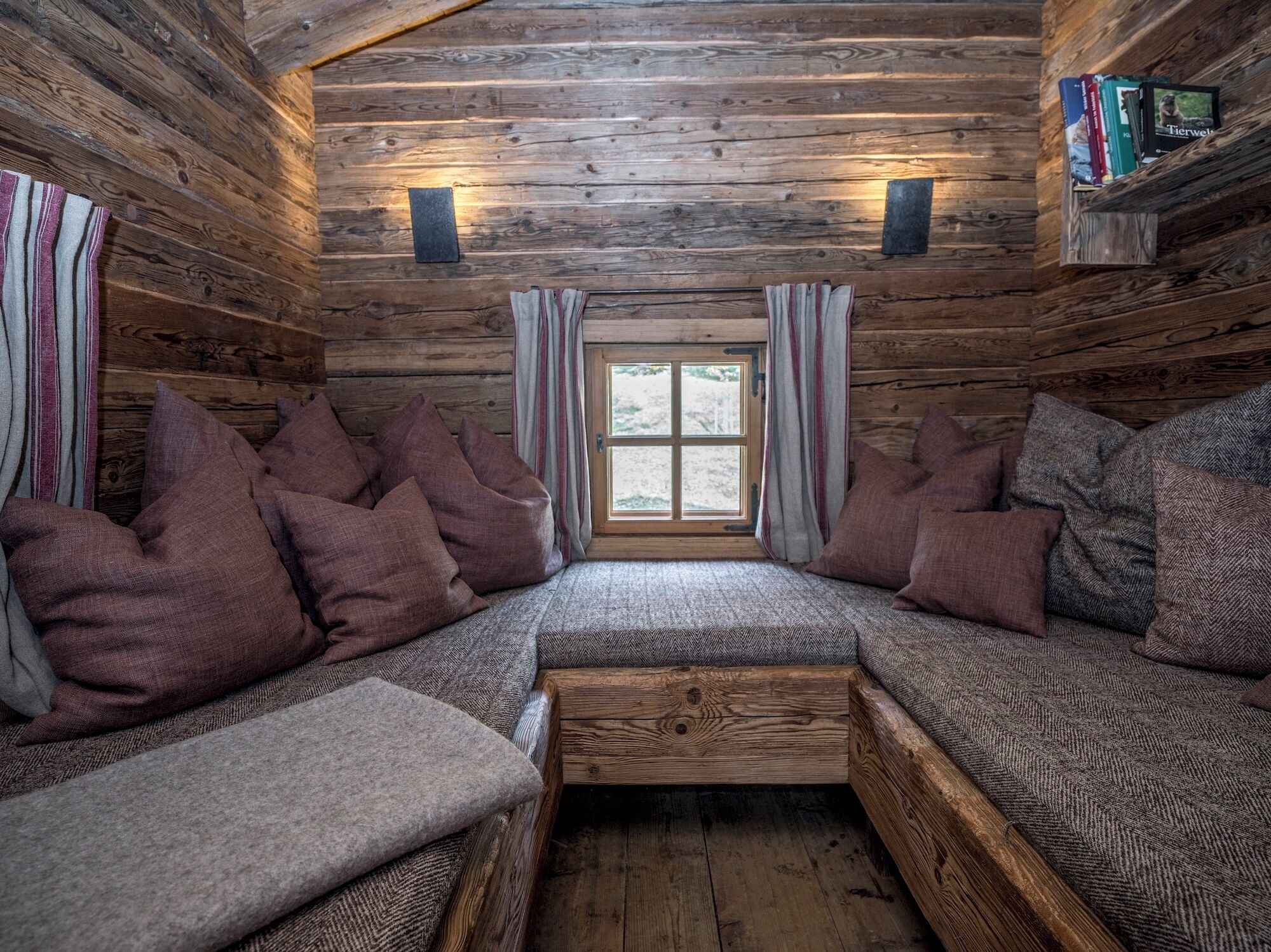 A cozy cabin interior with a wooden interior, a bench with cushions, and a bookshelf with books and a framed picture.