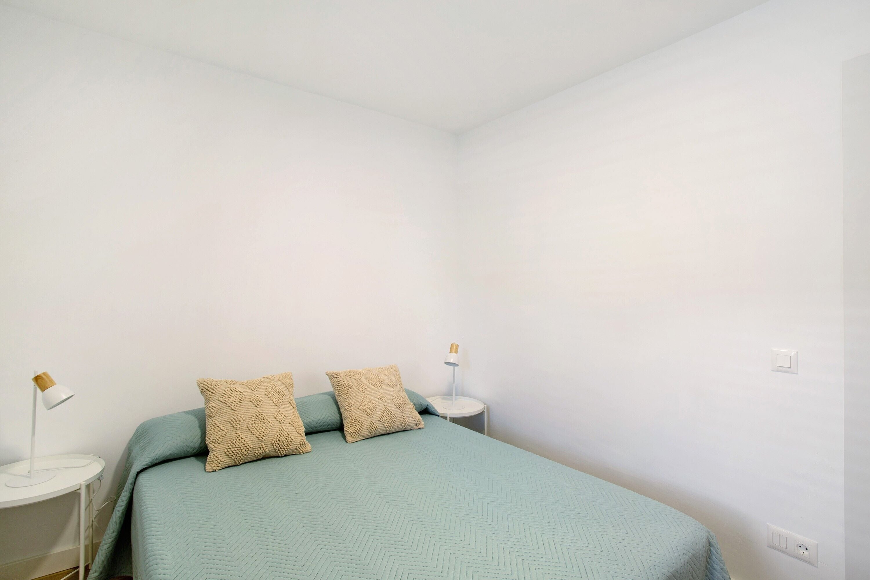A simple, clean room with a bed, two pillows, a side table with a lamp, and a wall socket.