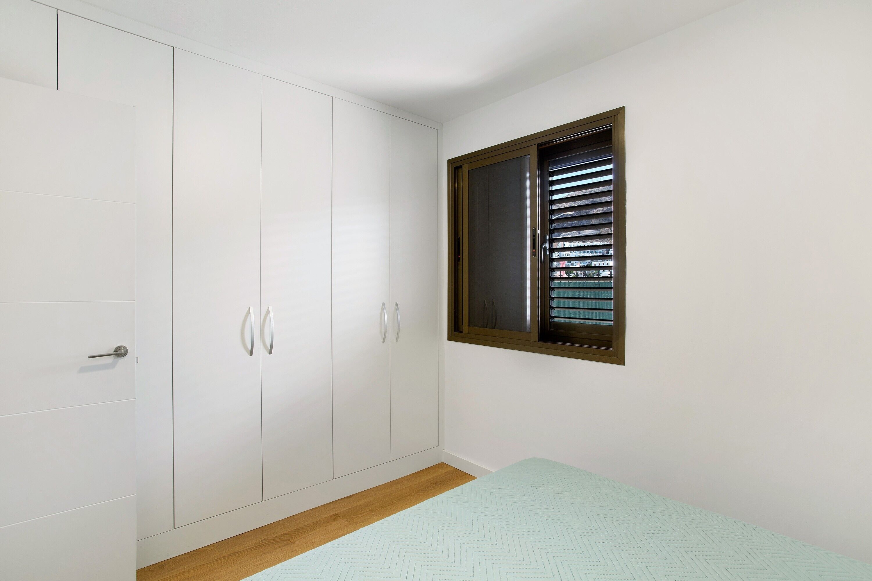 A bedroom with a wardrobe, a window with blinds, and a bed with a patterned bedspread.