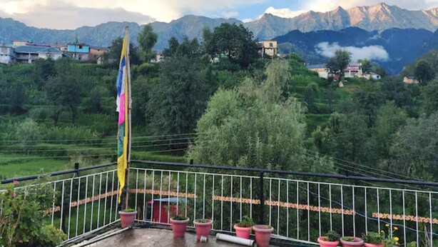 A balcony with potted plants and a flag, overlooking a mountainous landscape with houses and greenery. - Shiva Home Stay (Dharamshala)