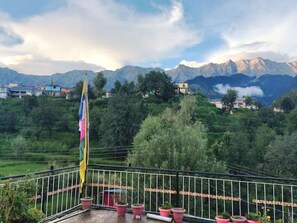 A balcony with potted plants and a flag, overlooking a mountainous landscape with houses and greenery. - Shiva Home Stay (Dharamshala)