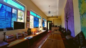 A spacious room with large windows, a long wooden desk, and a cozy seating area. - Shiva Home Stay (Dharamshala)